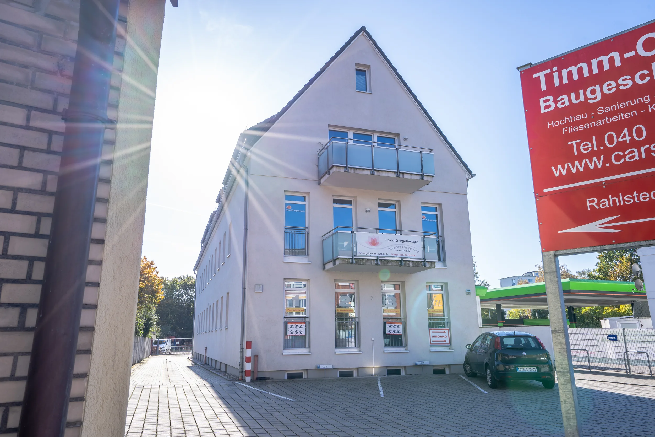 Weißes Wohn- und Geschäftshaus mit Satteldach und Balkonen, davor Parkplatz mit grünem Auto, rote Bauschild sichtbar
