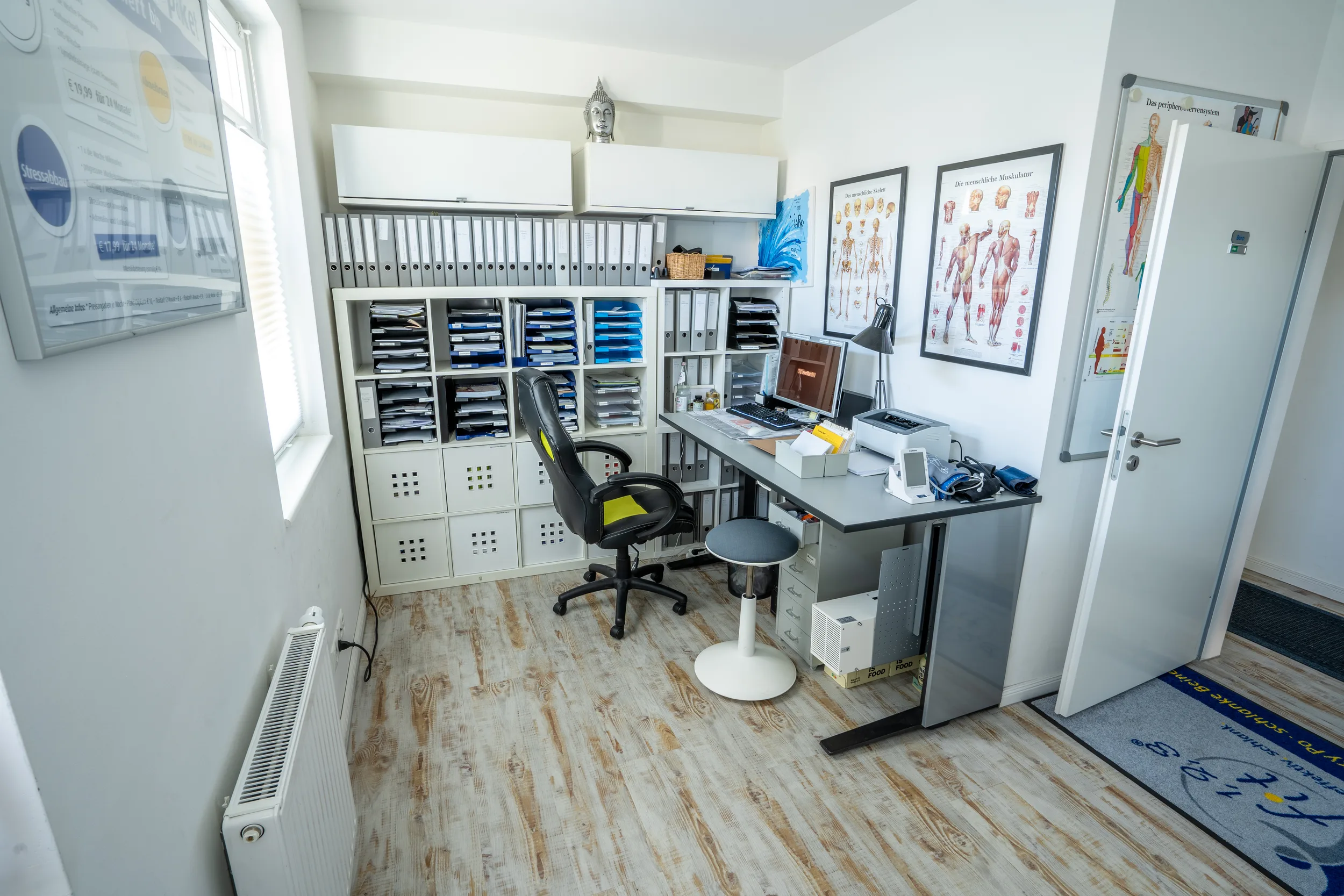 Helles Praxiszimmer mit Schreibtisch, Computer, schwarzem Behandlungsstuhl und Wandregalen mit Materialien
