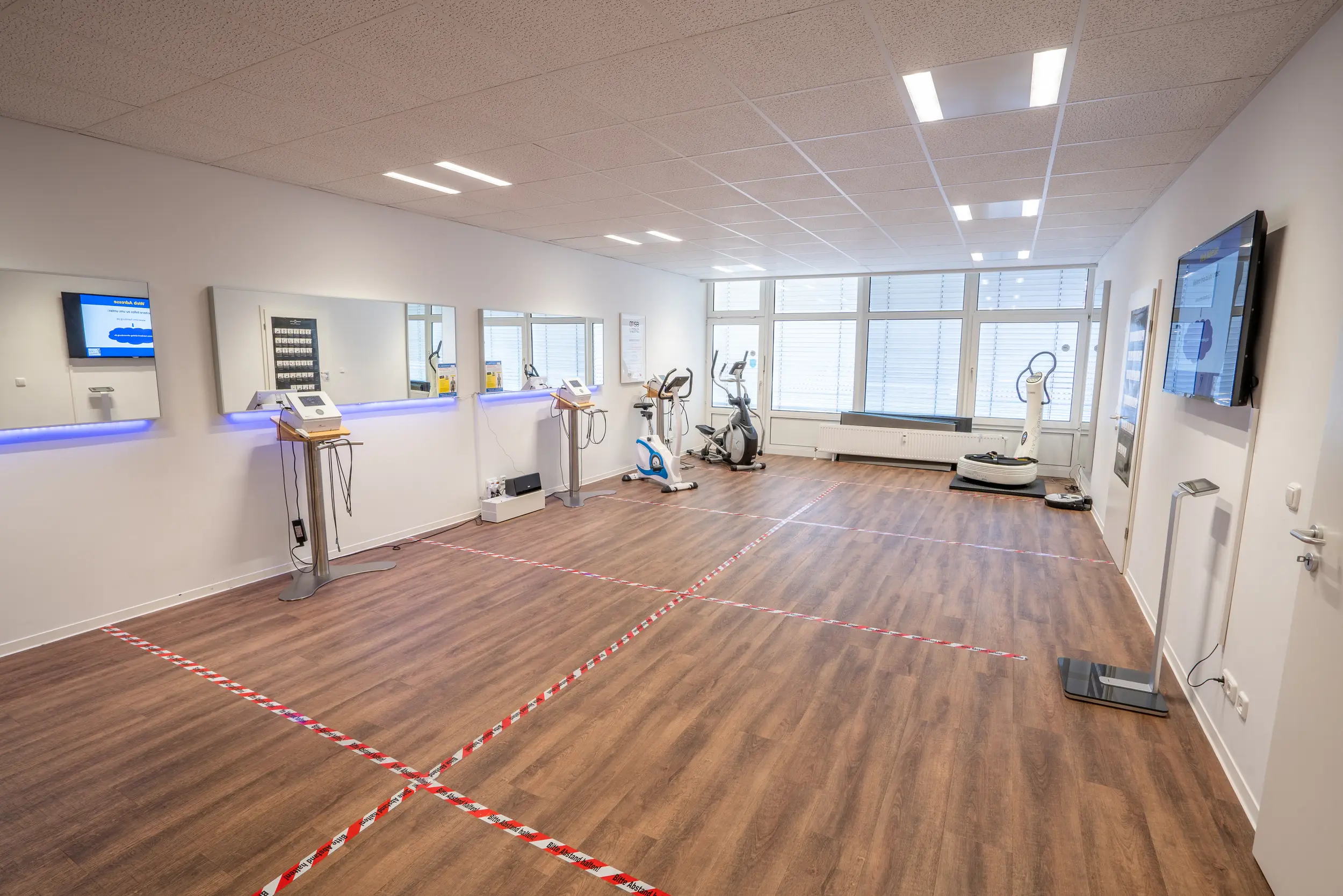 Modernes, helles Behandlungsstudio mit Holzparkettboden, Empfangstheke mit lila Akzentbeleuchtung links, mehrere medizinische Trainingsgeräte in der Mitte, große Fenster mit weißen Jalousien, Bildschirme an den Wänden, rote und weiße Absperrungstape auf dem Boden